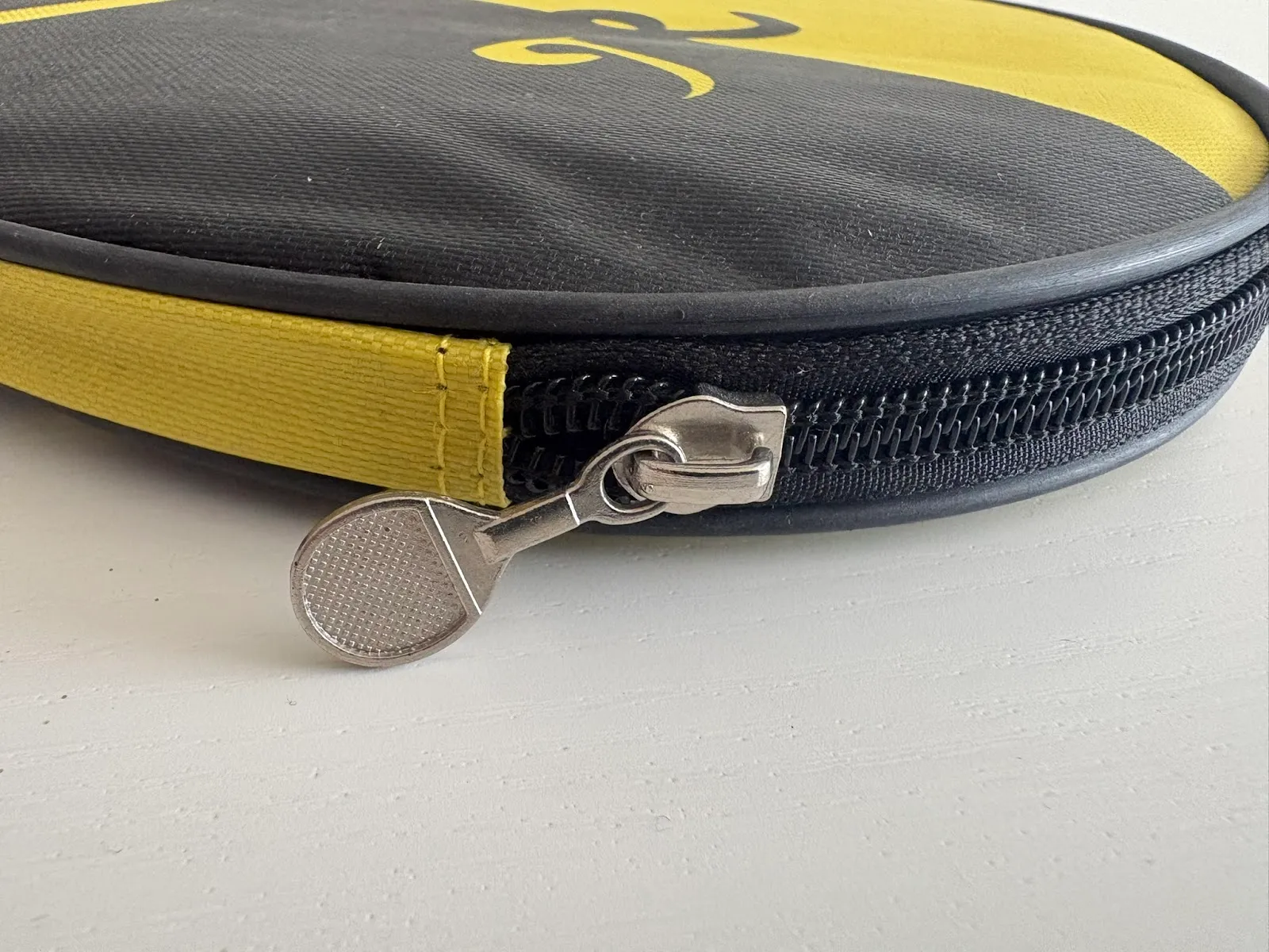 Close-up of a black and yellow round zippered case on a white surface, showing black zipper teeth, stitched yellow fabric edging and a silver metal zipper pull shaped like a small racket.