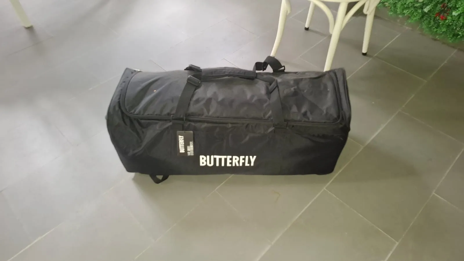 Black Butterfly travel bag with wheels and white Butterfly logo on tile floor next to a white chair.
