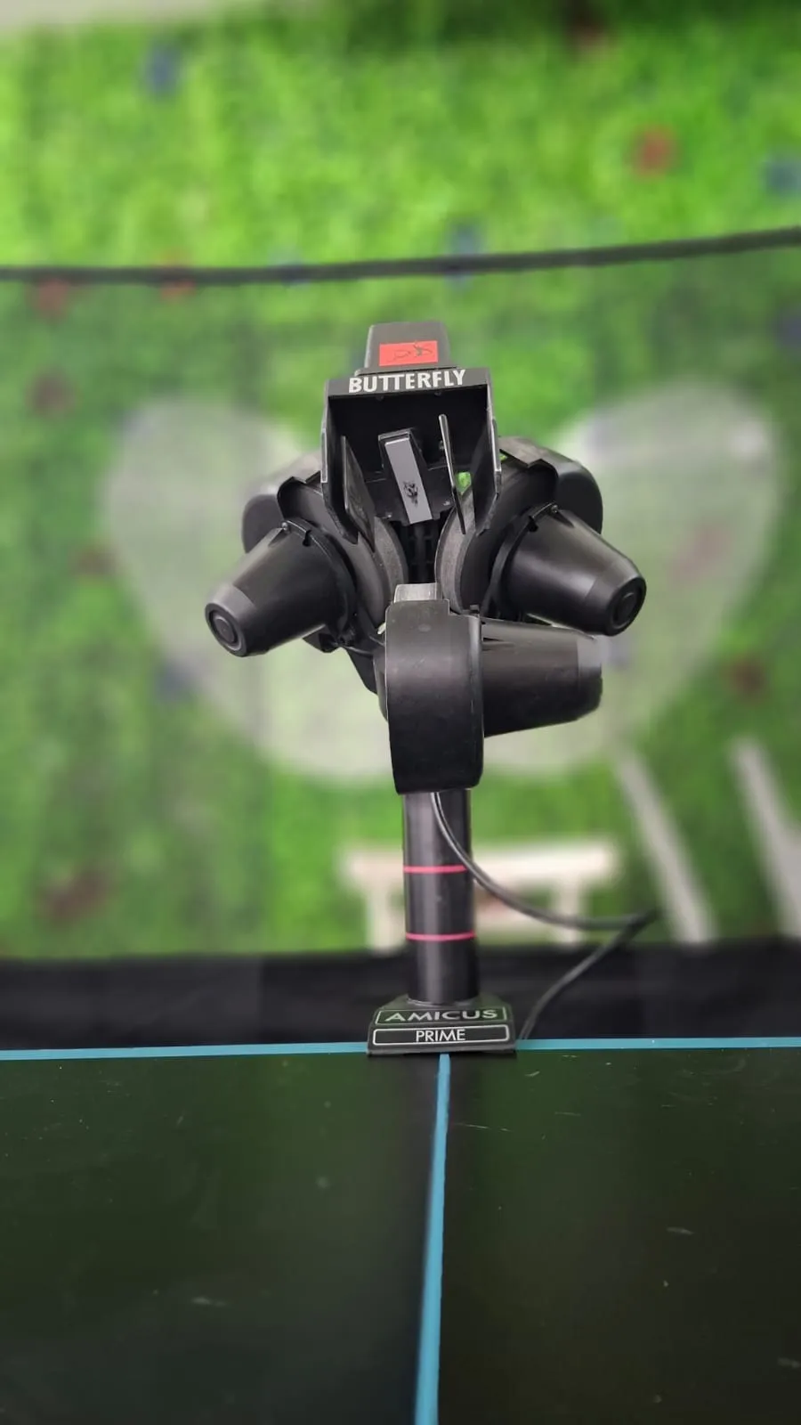Front-on view of the Butterfly Amicus Prime three-wheel projection head with red Butterfly logo and Amicus Prime base plate, mounted on a black table.