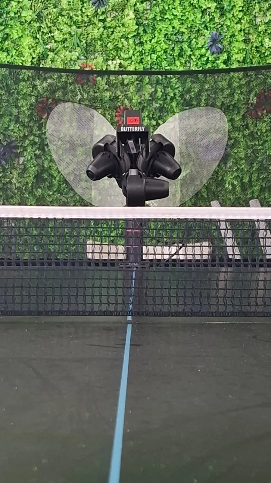 Butterfly Amicus Prime mounted on a black table behind the net, framed by a green leafy backdrop and the dark mesh of the recycling system.