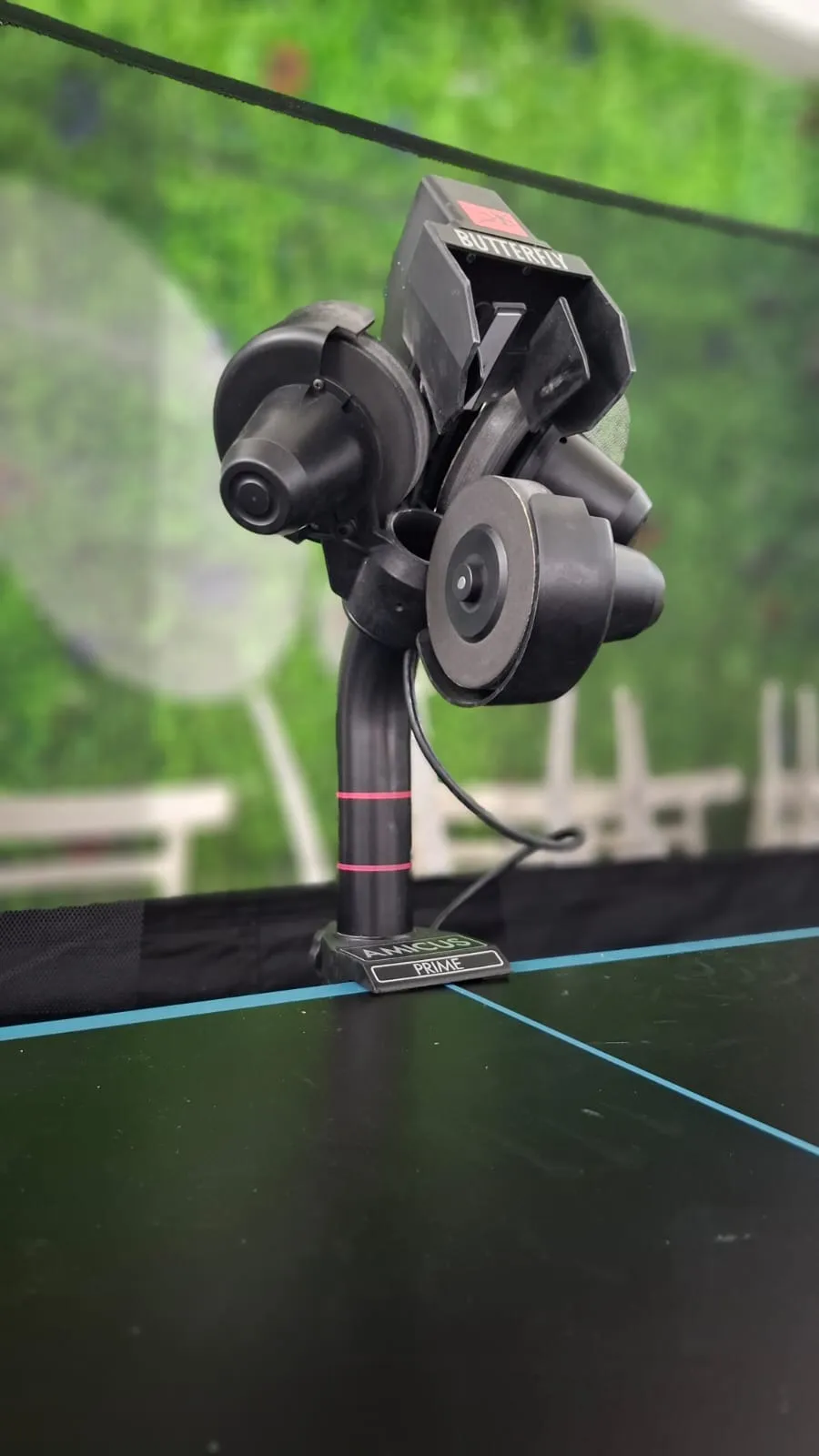 Side view of the Butterfly Amicus Prime mounted on the edge of a black table, showing the three projection wheels and the Amicus Prime logo on the base.