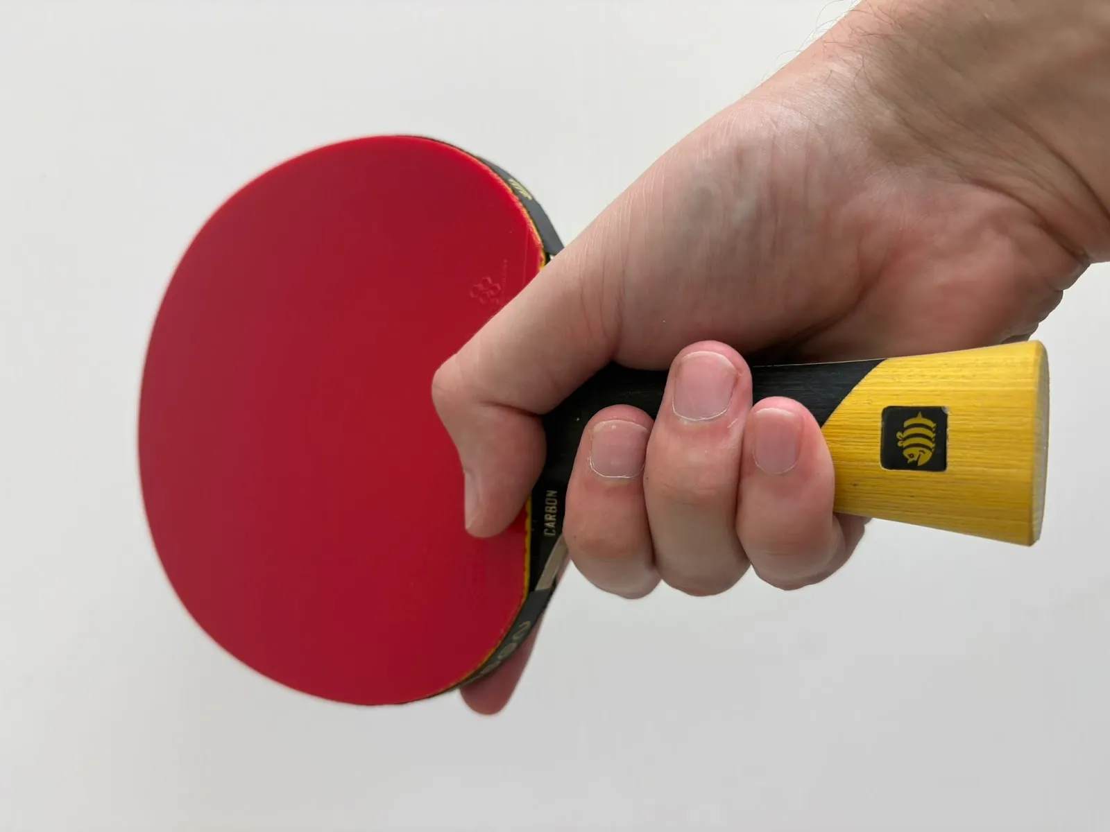 Close-up of a hand gripping a table tennis paddle with a red rubber face and a yellow wooden handle against a white background.
