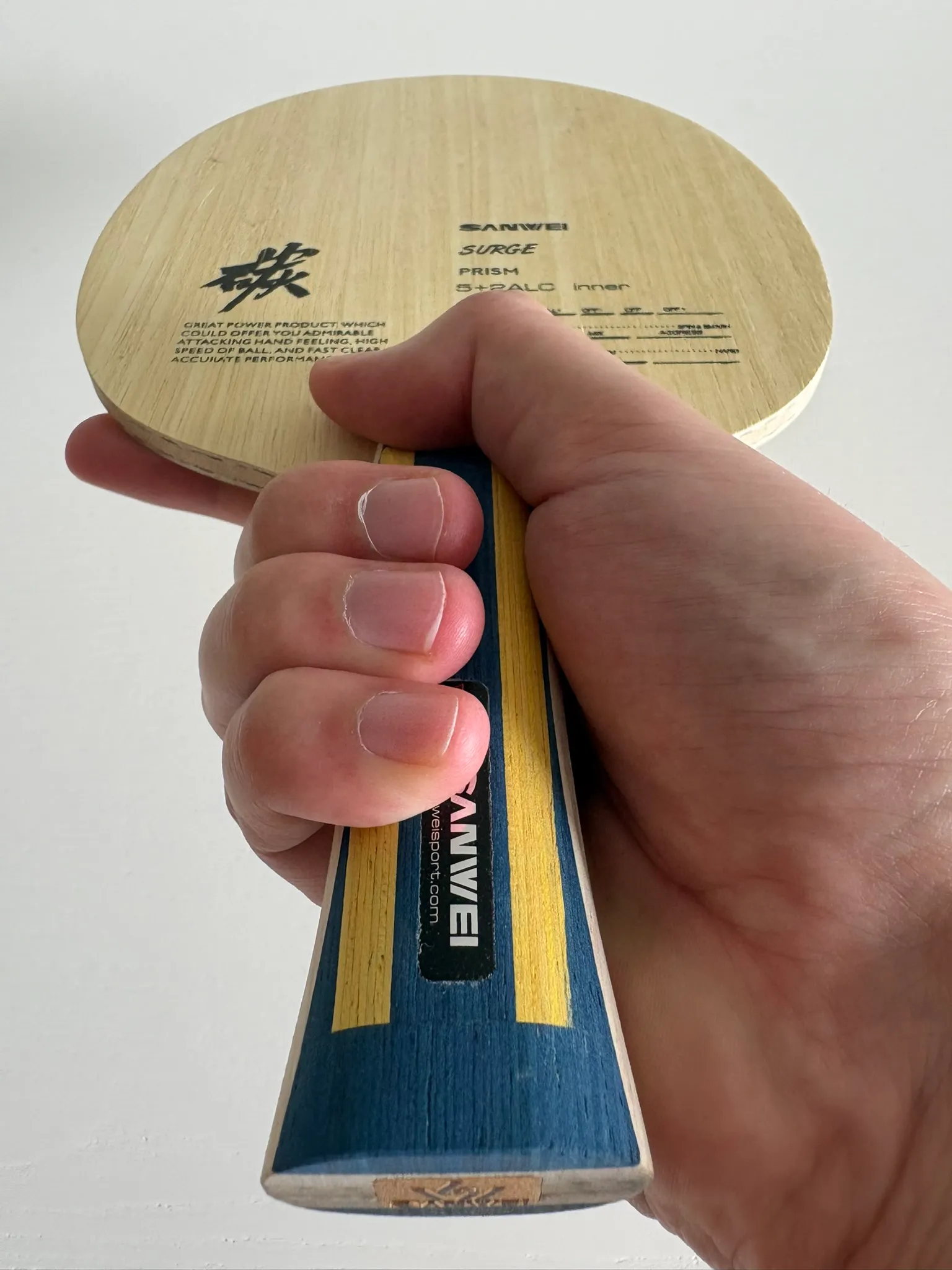Hand holding the bare Sanwei Surge Prism blade, showing the light wood face and the blue and yellow flared handle.