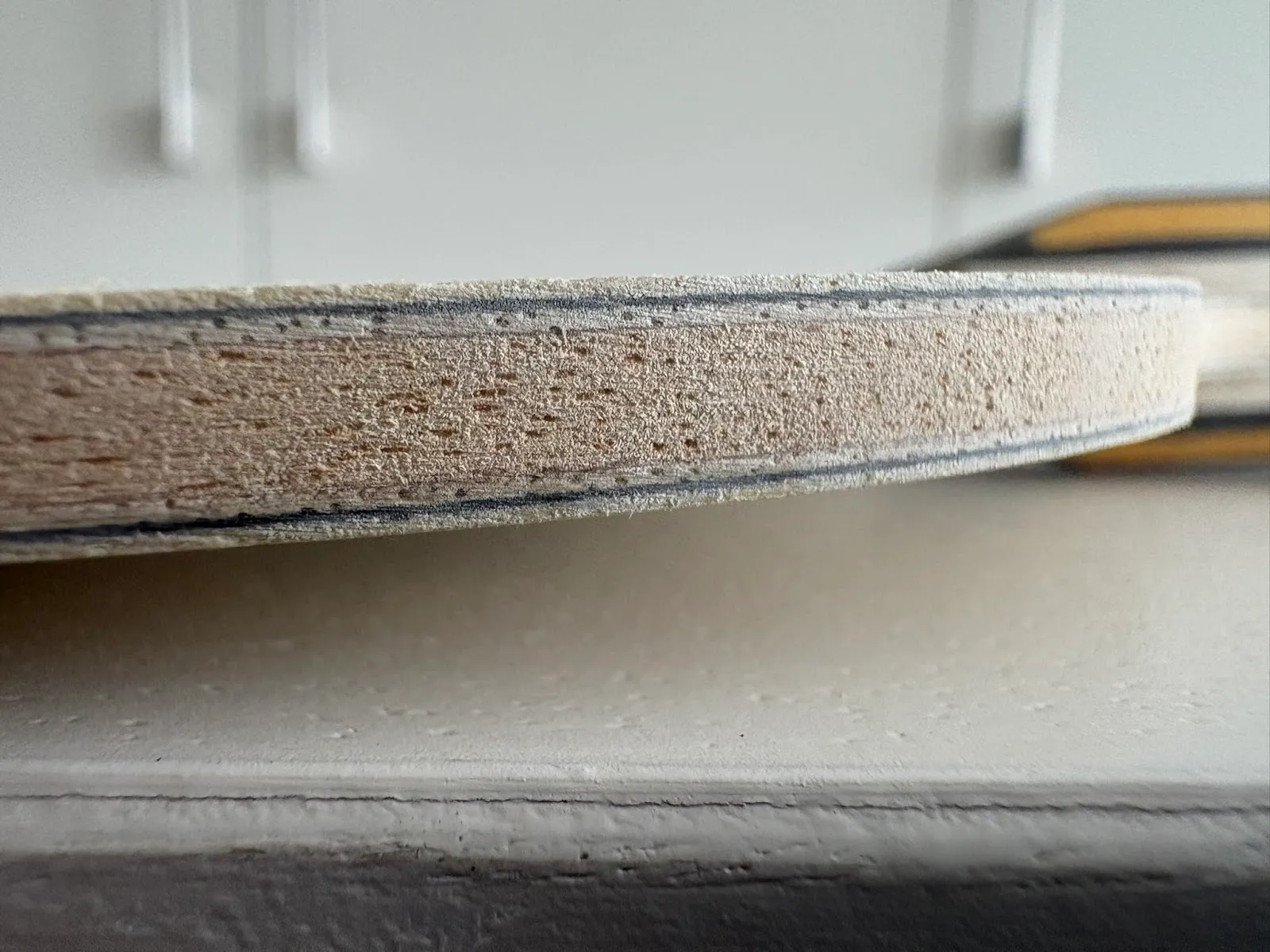 Close-up side view of a layered wooden edge showing porous wood texture and thin dark veneer lines, resting above a painted white surface with blurred cabinets in the background.