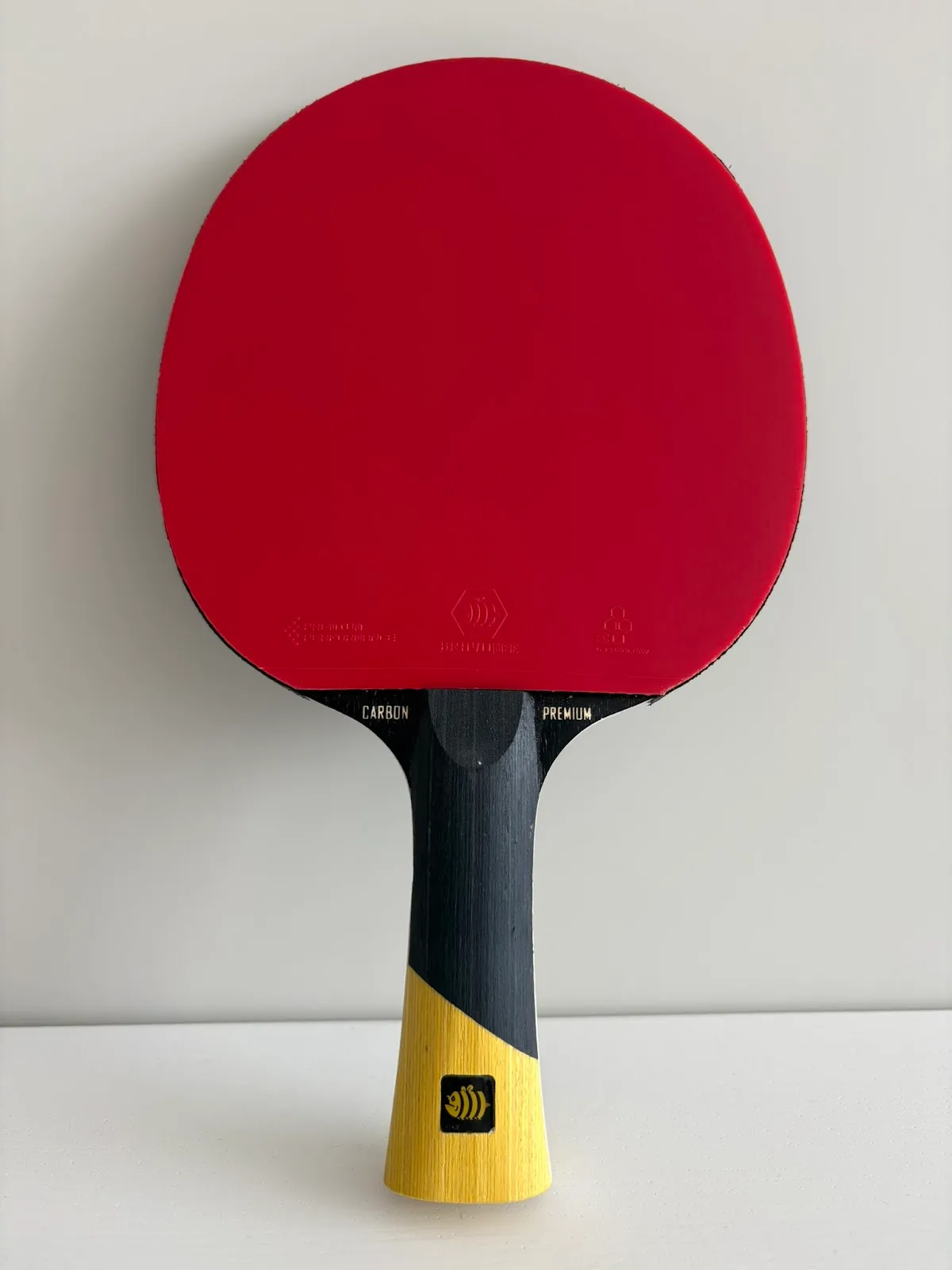 Table tennis paddle with a red rubber face and a black and yellow handle standing upright on a white surface against a pale gray background.