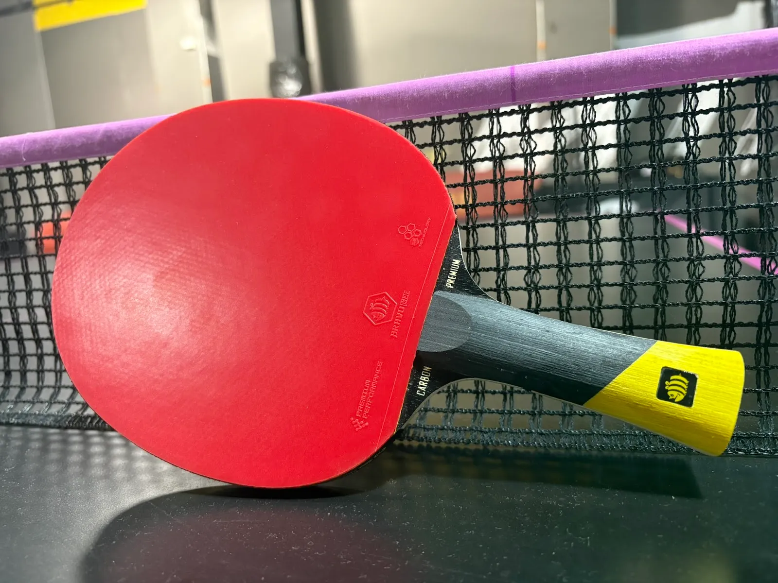 Close-up of a red table tennis paddle with a black and yellow handle resting against a black mesh net topped by a purple fabric band.