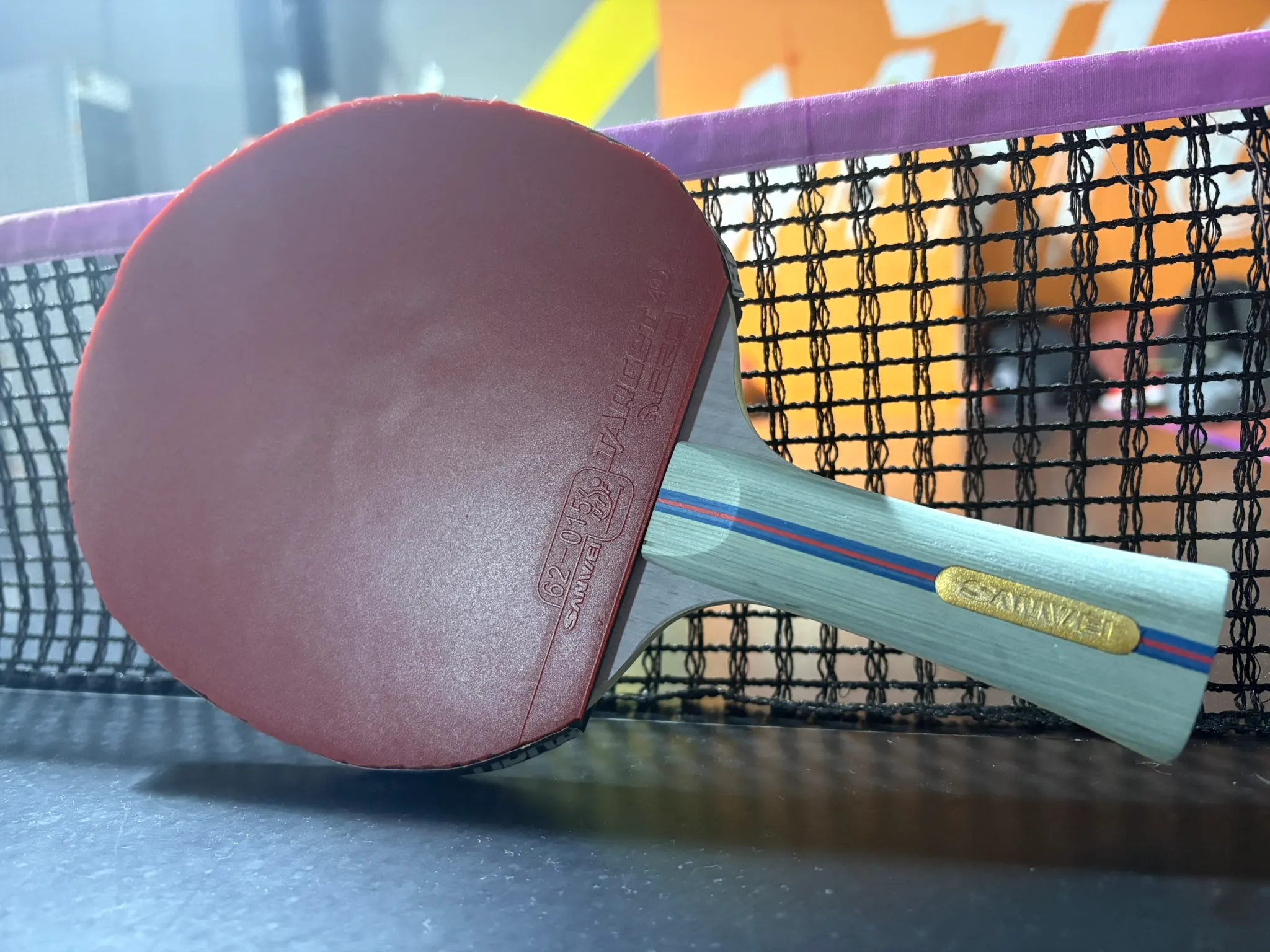 Table tennis blade with red rubber leaning against a purple-topped net on a dark table in a sports hall.