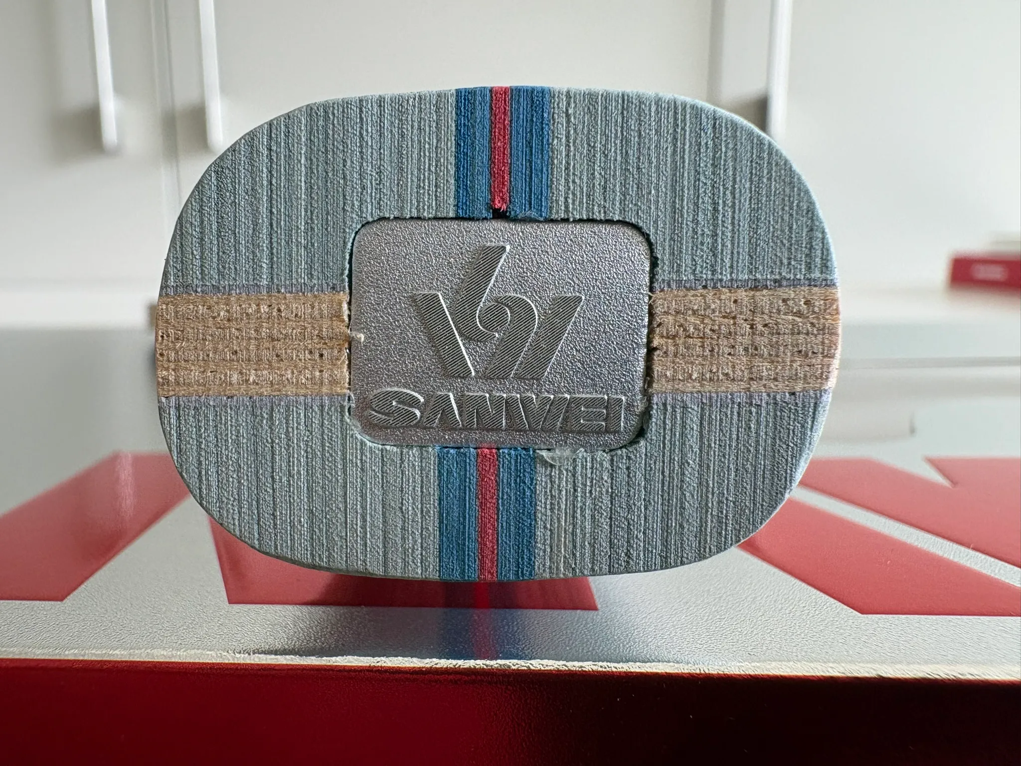 Close-up of the flat butt end of the handle showing layers of pale veneer, blue and red stripes, and an inset silver Sanwei badge.