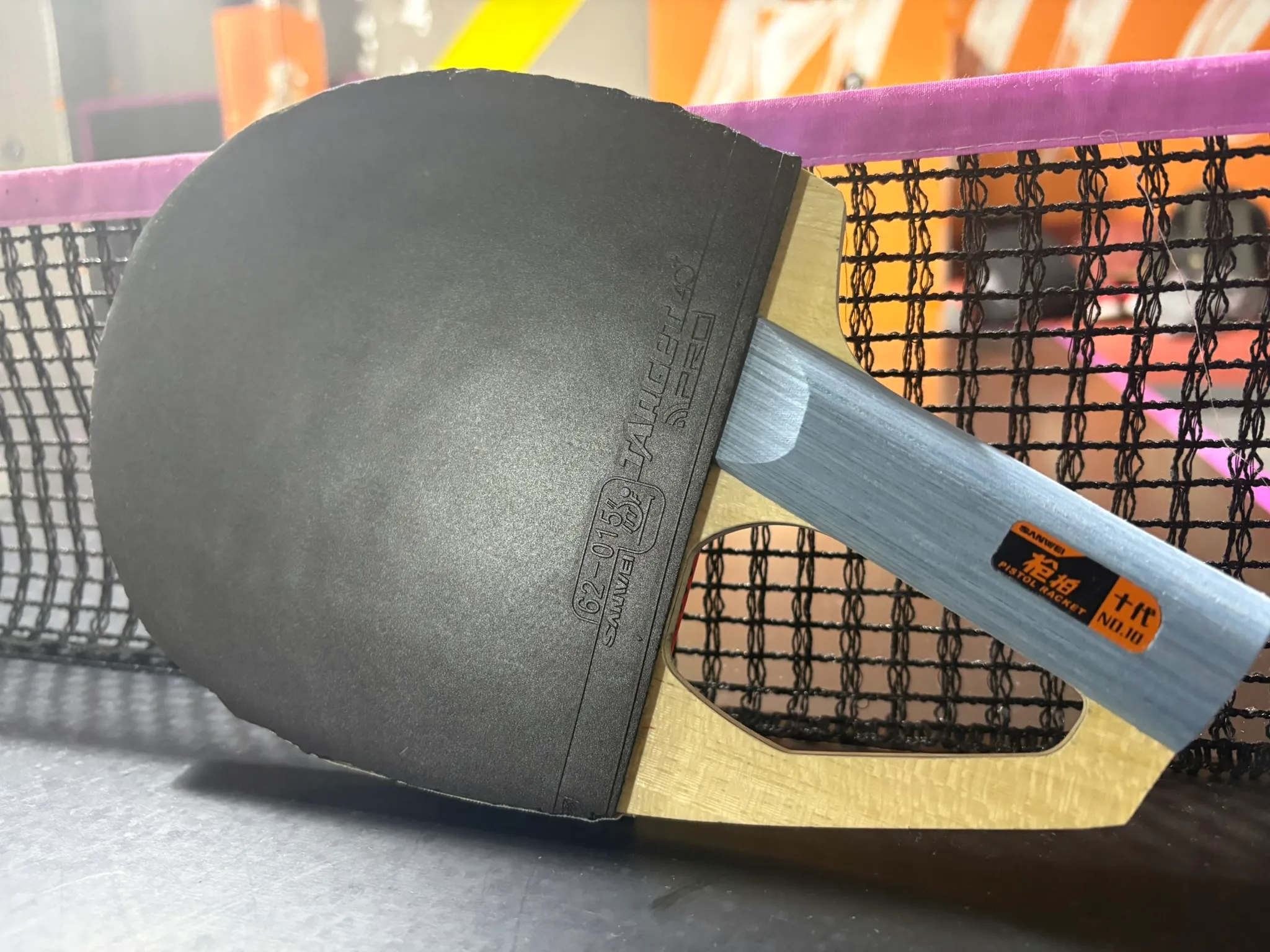 Assembled Sanwei Pistol Racket with black rubber resting against a table tennis net.