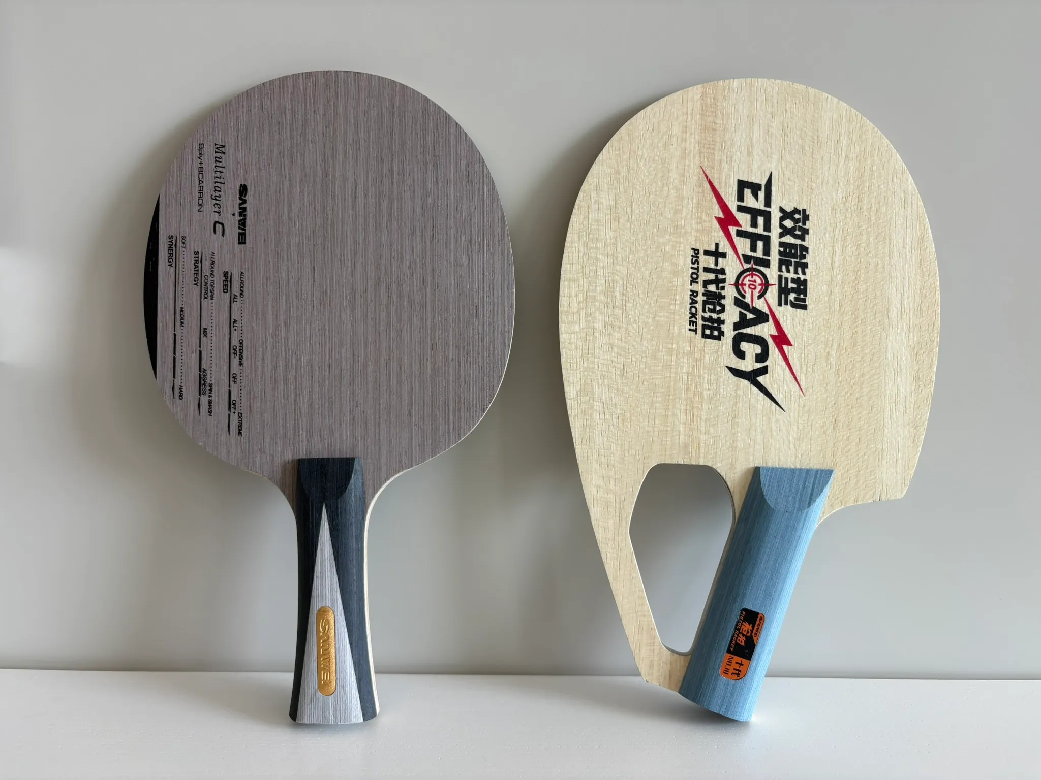 Sanwei Pistol Racket beside a conventional shakehand blade on a white surface.