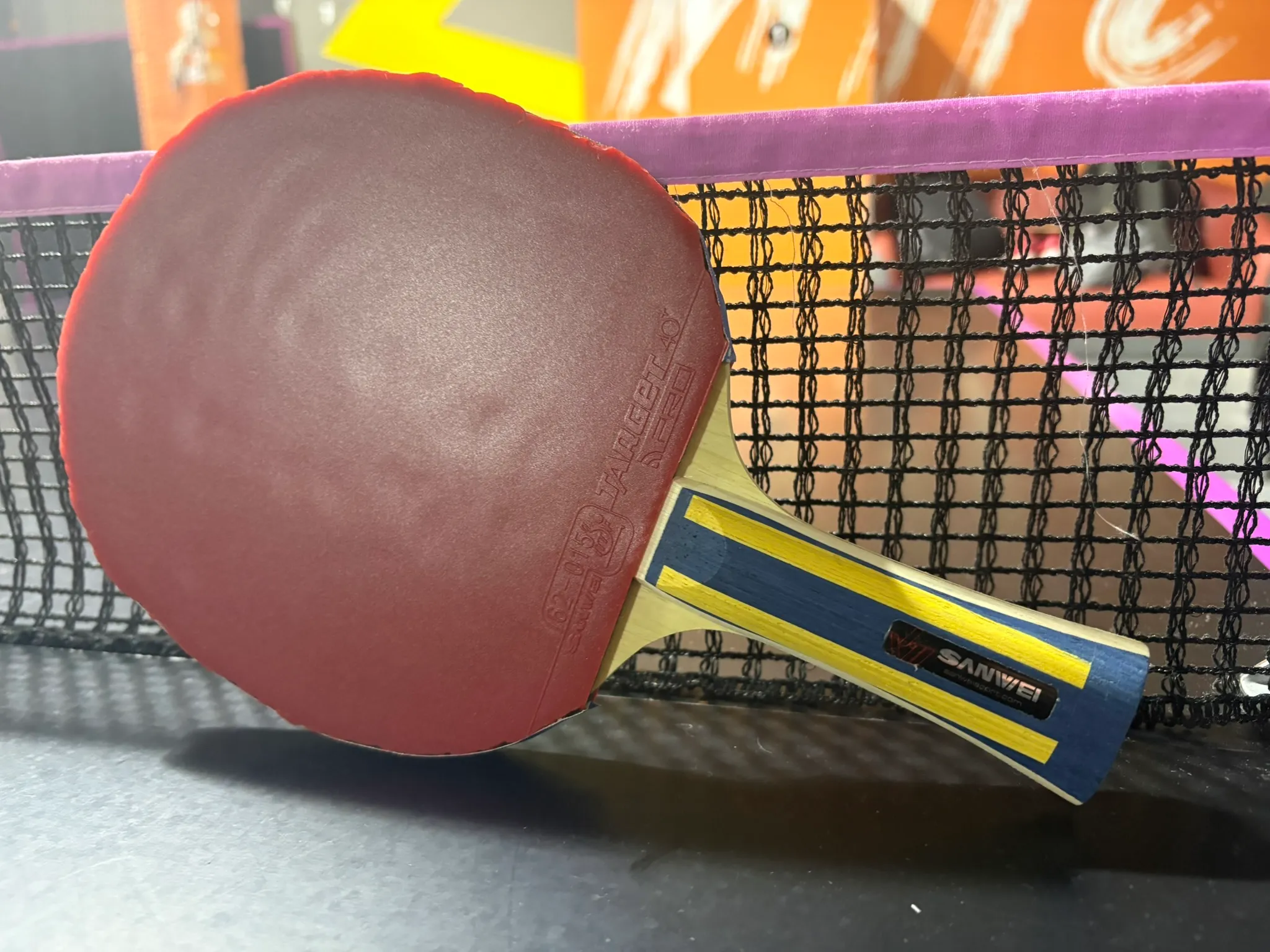 Assembled Sanwei Surge Prism racket with red rubber resting against a table tennis net.
