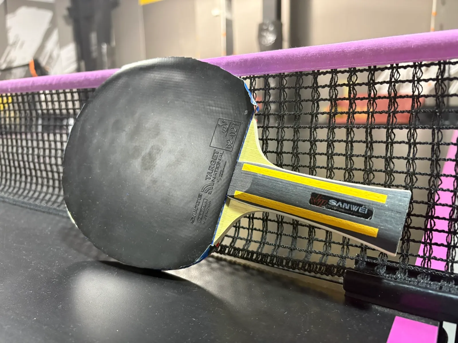 Close-up of a table tennis paddle with black rubber face and a wooden handle labeled "SANWEI" resting against a woven ping-pong net with a purple top band on a dark table surface.