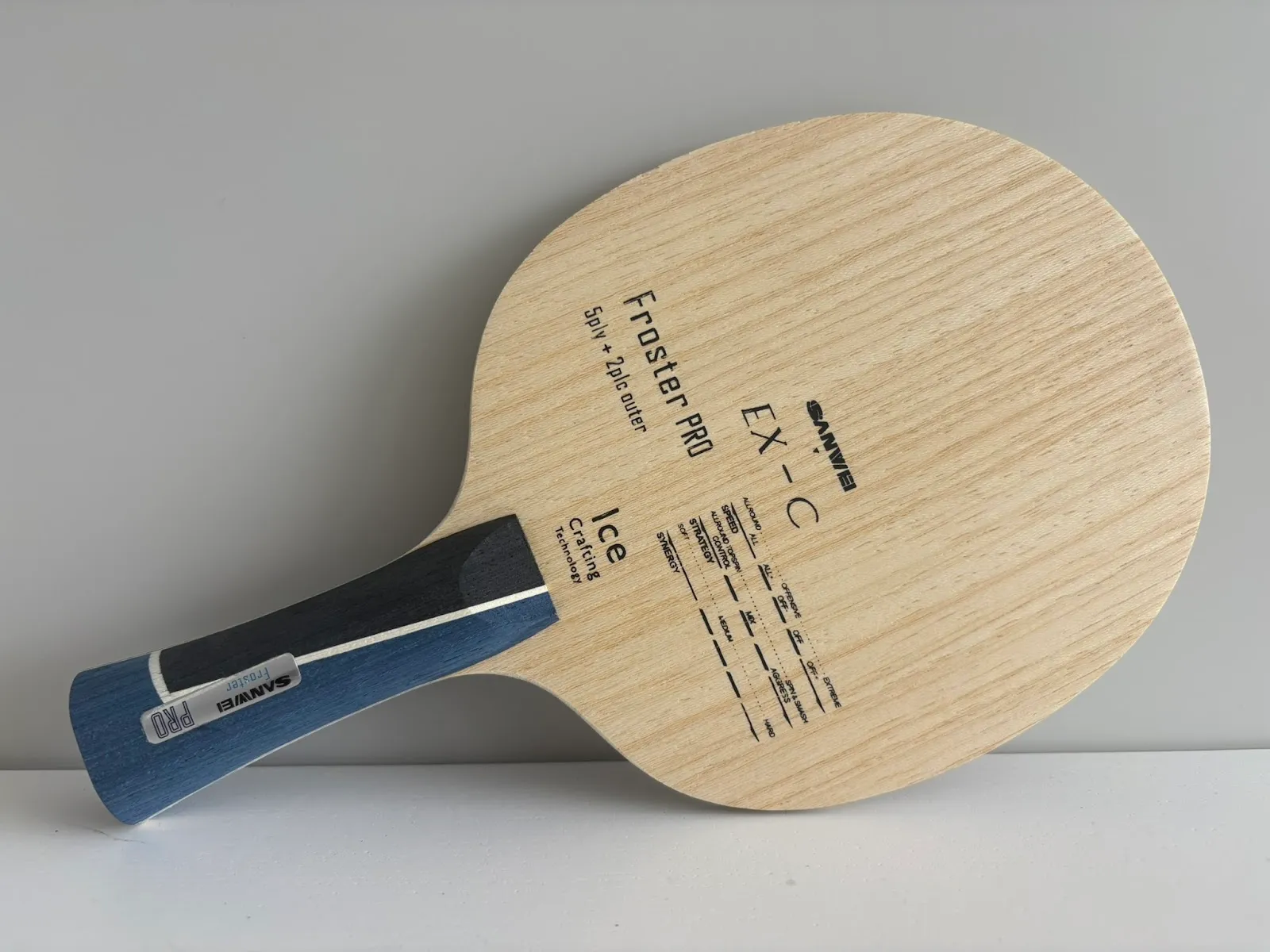 Angled close-up of a light-wood table tennis blade with a blue and black flared handle, showing printed markings including 'EX-C', 'Froster PRO', and 'Ice', on a grey surface.