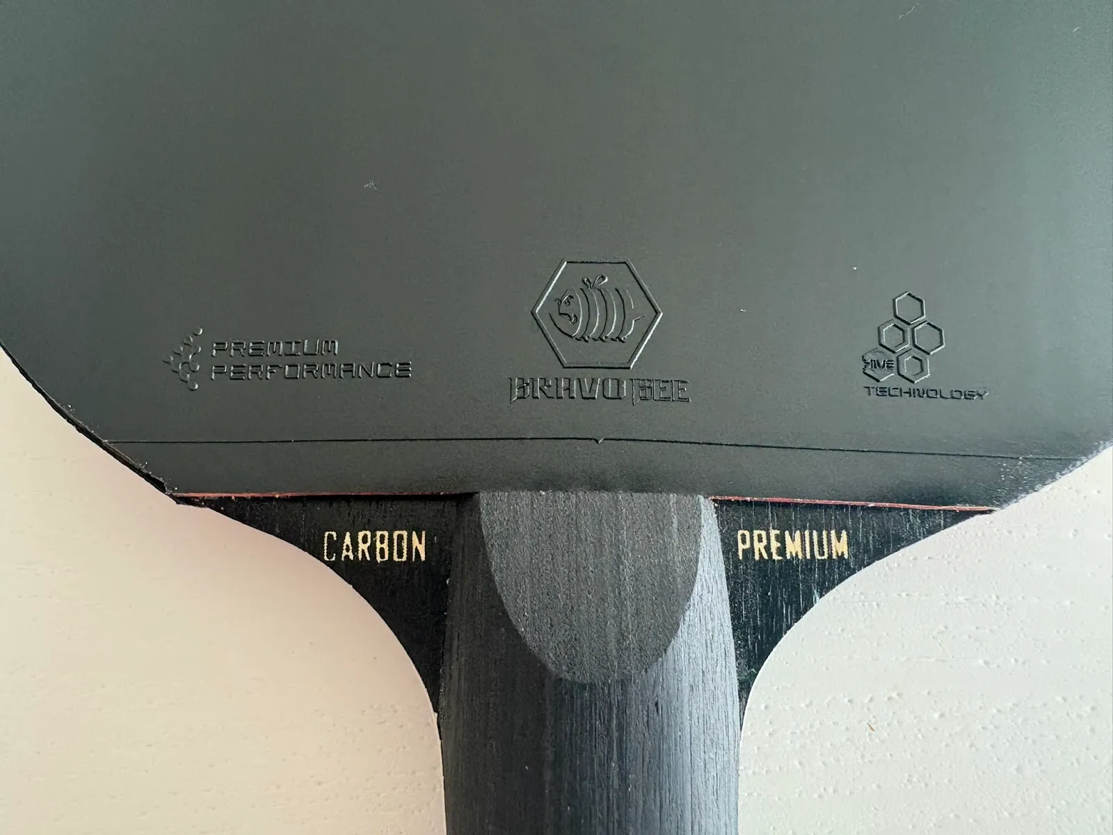 Close-up of a black table tennis paddle top with embossed logos on the rubber and a dark wooden handle showing gold text 'CARBON' and 'PREMIUM'.
