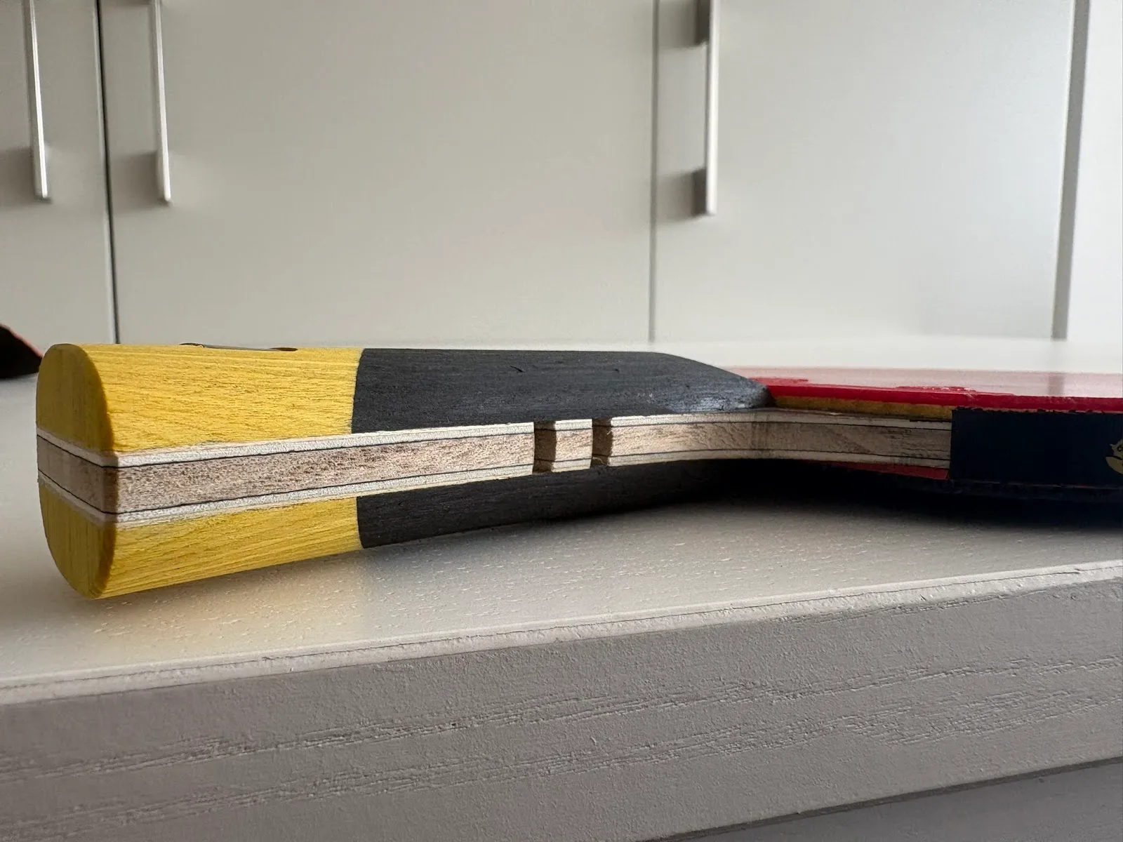 Close-up of a table tennis paddle handle and blade edge showing laminated wood layers, yellow and black painted handle sections and red rubber surface, resting on a white surface.