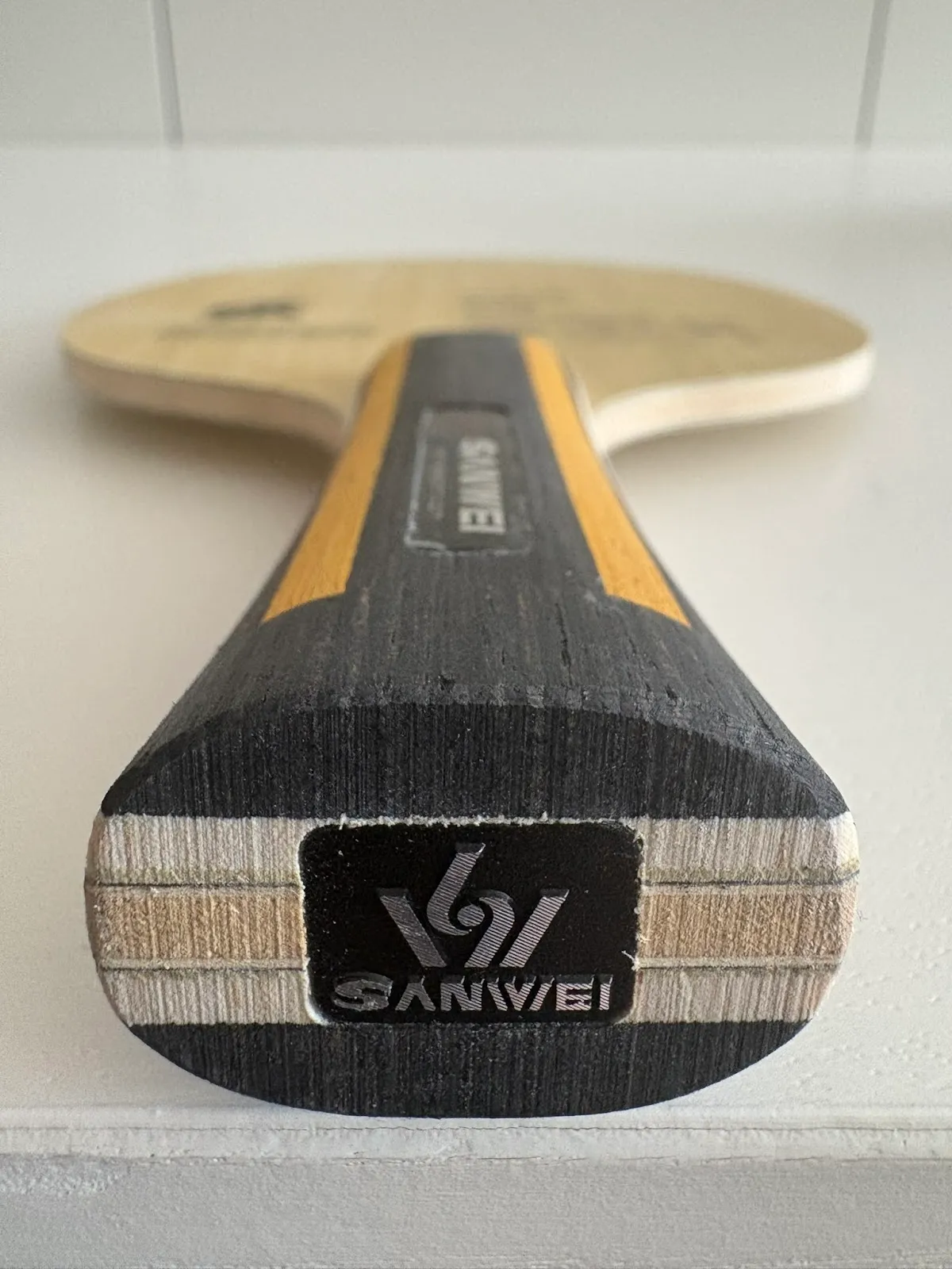 Close-up of the end of a wooden table tennis paddle handle showing layered wood grain and a black rectangular badge with the SANWEI logo, with the paddle blade blurred in the background.