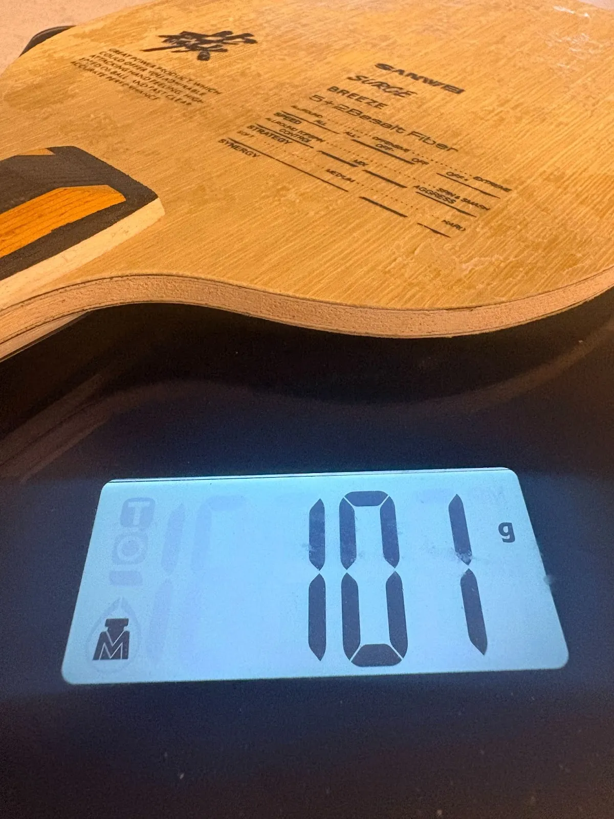 Layered wooden board with printed text and a small black-and-yellow inlay resting on a digital scale display reading 101 g